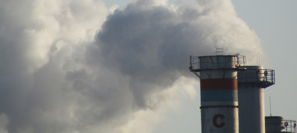 Industrial chimneys release thick plumes of smoke into the atmosphere, illustrating the persistent challenge of regulating stationary emission sources to mitigate the global respiratory health crisis.