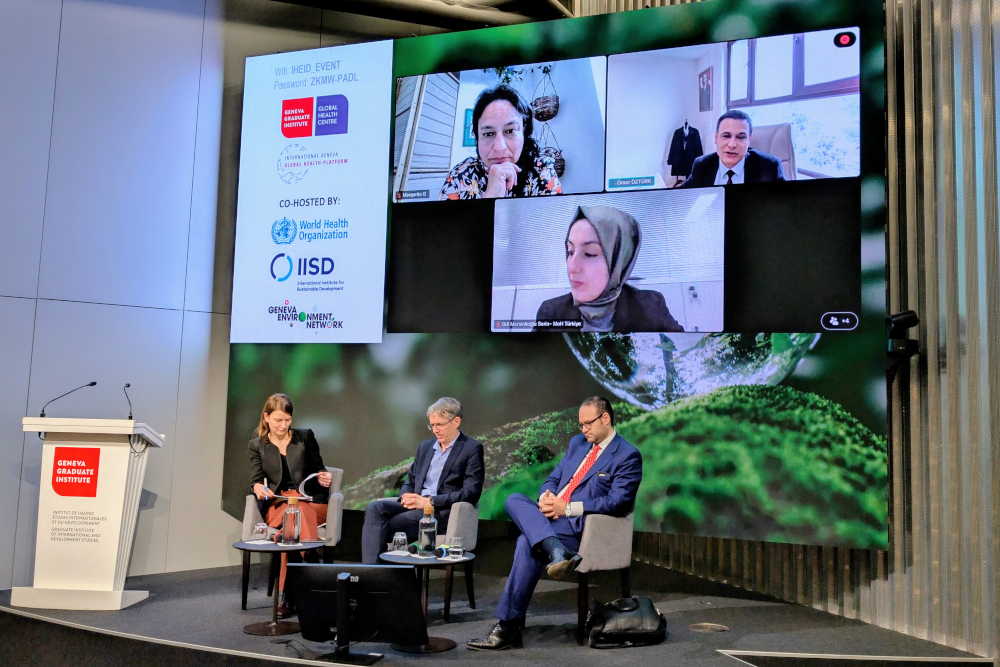 Panellists Diarmid Campbell-Lendrum (WHO, stage centre), Miguel Ruiz Botero (Colombian UN Mission, stage right), Margarita Gutierrez (IISD, screen left), Ömer Öztürk (Türkiye Min. of Environment, screen right), and Gül Mersinlioğlu Serin (Türkiye Min. of Health, screen centre) discussing two-speed multilateralism in Geneva.