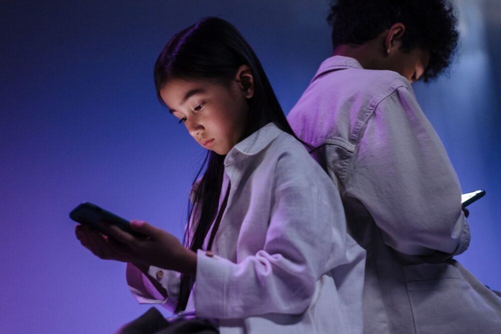 Two children engrossed in using their smartphones in a dimly lit setting. A lack of digital governance exposes young people to algorithmic harms and severe health risks, warn WHO and France.